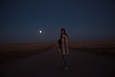 Woman wrapped in blanket standing on street amidst field against sky at dusk