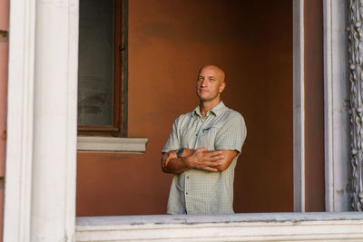 A balding middle-aged man in a plaid shirt stands against a painted wall