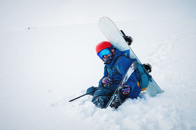 Man with ski equipment and a snowboard is sitting a snowy mountain. blue sky and snowy mountain in