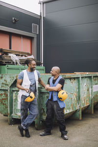 Smiling male colleagues with hardhat looking at each other against against metal container