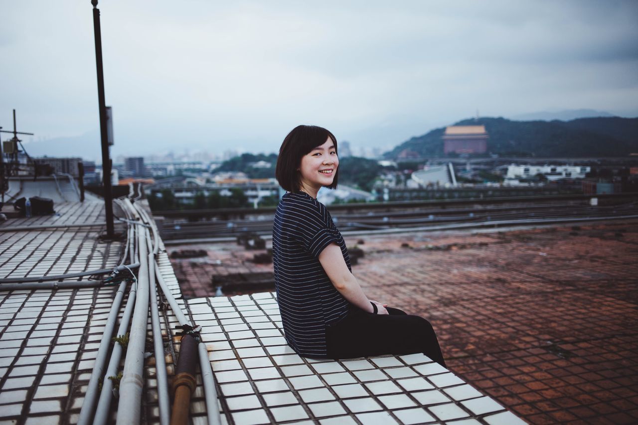 architecture, one person, building exterior, built structure, sky, lifestyles, real people, young adult, city, leisure activity, side view, casual clothing, adult, sitting, nature, women, young women, day, outdoors, beautiful woman, cityscape, hairstyle, contemplation