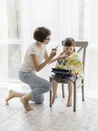 Mother cuts her son's hair by herself. little boy sits and holds digital tablet. new normal