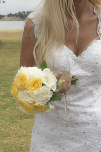 Midsection of bride holding flower bouquet and champagne flute on field