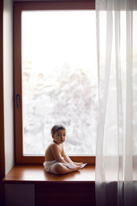 Baby boy in bath towel after washing sitting on the windowsill at the big window in winter