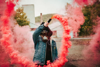 Person in gas mask and hood keeping bomb with pink fume in raised hand in countryside person