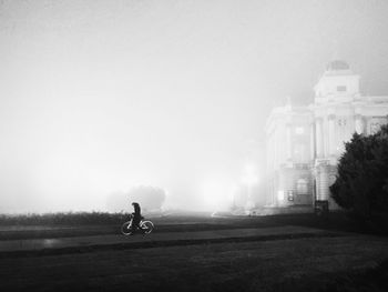 Person riding bicycle by building against sky during foggy weather