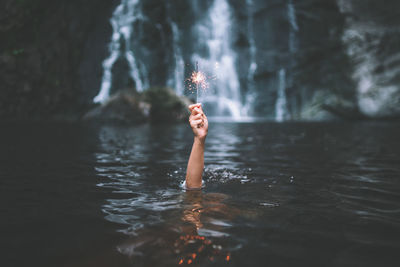 Cropped hand holding sparkler against waterfall in lake