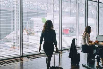 Rear view of businesswoman walking with luggage by female colleague sitting at airport departure area