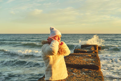 A woman with blond hair in a hat and a fur coat walks along the sea along a stone breakwater