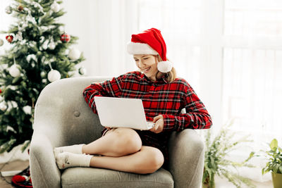 Happy teenage girl wearing santa claus hat using laptop in christmas room decorated