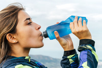 Side view of positive young ethnic female athlete in sportswear smiling and looking at camera while drinking water after workout in mountains
