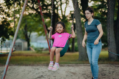 Cute girl sitting on swing while mother standing at playground