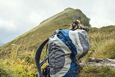 Large gray blue hiking backpack, trekking poles among green plants on background of  mountain peak