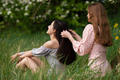 Two young girls in dresses are sitting under a white tree and braiding each other's hair