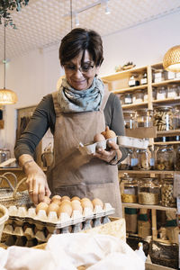 Positive elderly woman with short dark hair in casual clothes standing and putting fresh chicken eggs on cardboard tray in eco friendly shop