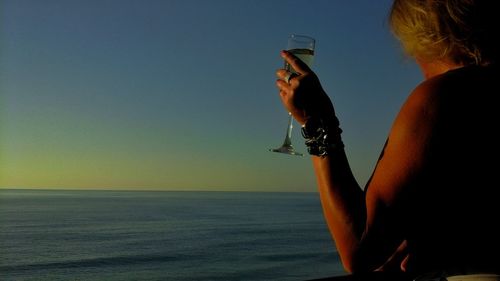 Shirtless woman holding champagne flute against sea and sky