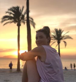 Portrait of woman smiling while sitting at beach against orange sky