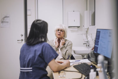 Senior woman explaining throat problem to otolaryngologist sitting at desk in medical clinic