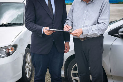 Midsection of man signing on paper with businessman against cars