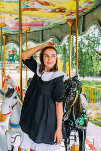 Beautiful brunette girl in a black dress poses on a bright carousel with horses