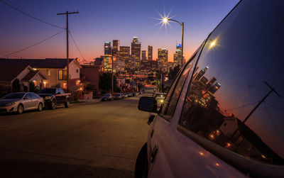 Illuminated street amidst buildings against sky during sunset