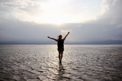 Rear view of silhouette mid adult woman with arms raised standing in sea against cloudy sky during sunset