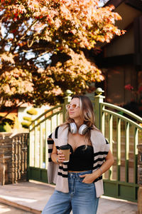 A woman wearing headphones drinks coffee and walks down the street in the countryside in autumn