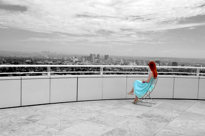 Woman looking at city while sitting on chair in building terrace against sky