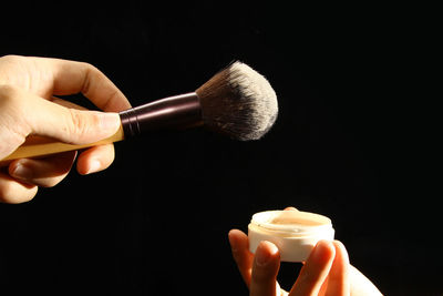 Cropped hands of person holding beauty product and make-up brush against black background