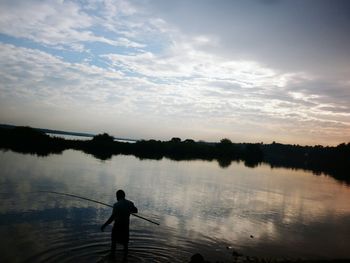 Silhouette boy standing in lake against sky during sunset
