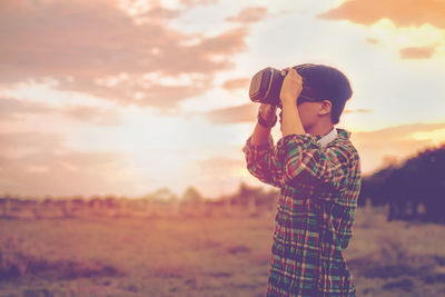 Boy photographing on field against sky during sunset