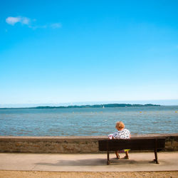 Woman sitting on bench at beach against blue sky