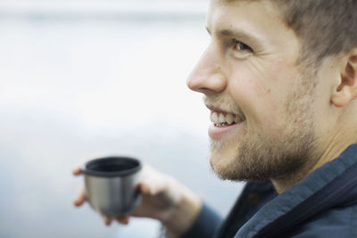 Happy man holding coffee cup against lake