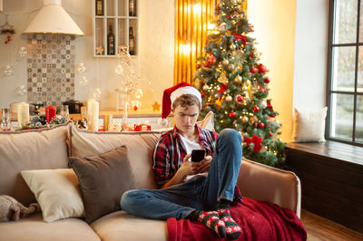 Teenage boy sits on the couch wearing a santa hat and with a phone in his hands. 