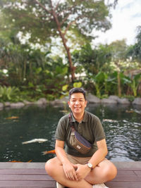 Portrait of smiling young man sitting on lake against trees