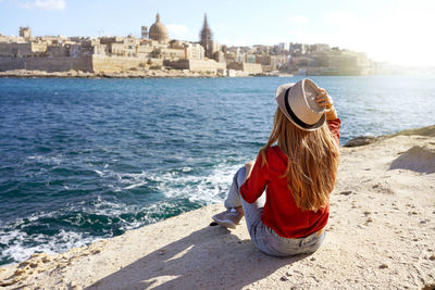 Woman with hat sitting on stone by sea looking at stunning panoramic view of valletta city in malta