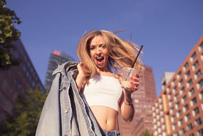 Low angle portrait of woman holding disposable cup against buildings