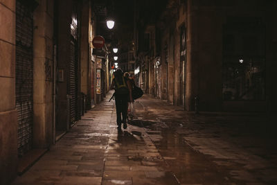 Rear view of woman walking on wet street in illuminated building