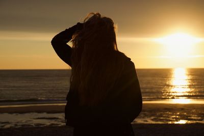 Rear view of woman with hand in hair standing at beach during sunset