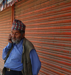 Senior man wearing dhaka topi listening to mobile phone against shutters