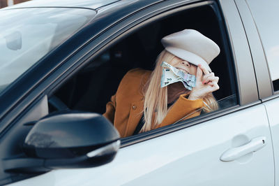 Middle aged woman sitting in a car and holding euro banknotes in her teeth, cost of living concept