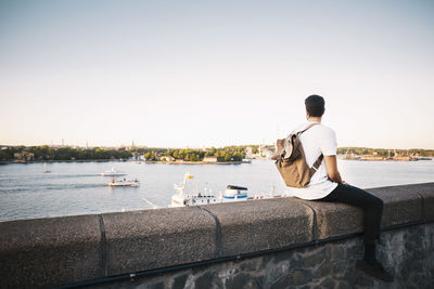 Male tourist looking at view while sitting on retaining wall of bridge against clear sky