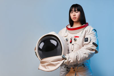 Young asian woman in spacesuit holding helmet and looking at camera under blue light before mission against gray background