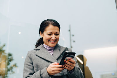 Low angle view of smiling businesswoman using smart phone while standing against building in city