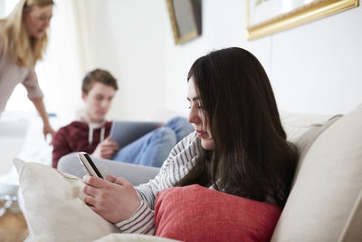 Young woman using mobile phone while mother looking at son with laptop in living room