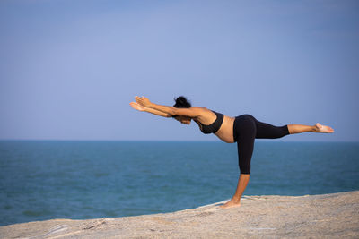 Full length of young woman exercising yoga on rock against sea against sky