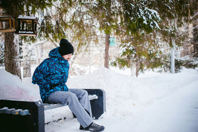 Young man sitting on bench against trees during winter
