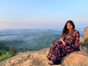 Portrait of young woman sitting on rock against sky