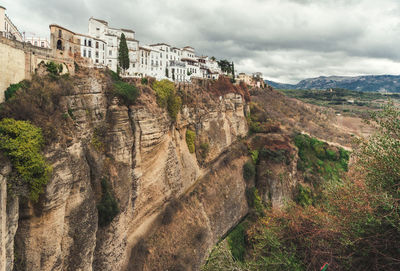 Buildings on rocky cliff against cloudy sky