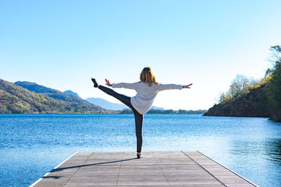 Rear view of woman doing yoga on jetty over lake against sky